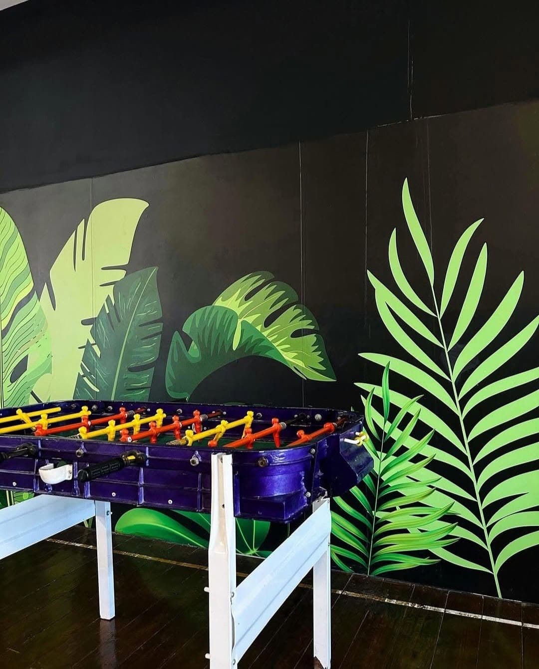 El metegol en el sector de juegos, contra la pared negra pintada con hojas de palmera verdes.
